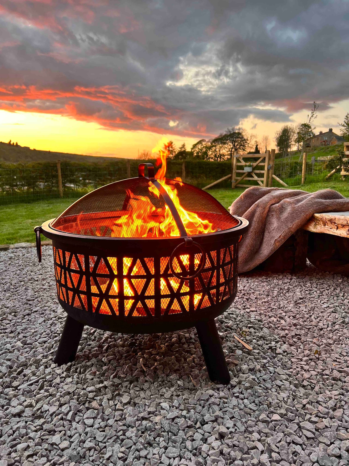 A fire pit is positioned on a gravel surface, with flames dancing inside. The background features a picturesque sunset casting warm hues over the landscape. A cozy blanket is draped nearby, suggesting a comfortable outdoor seating arrangement.