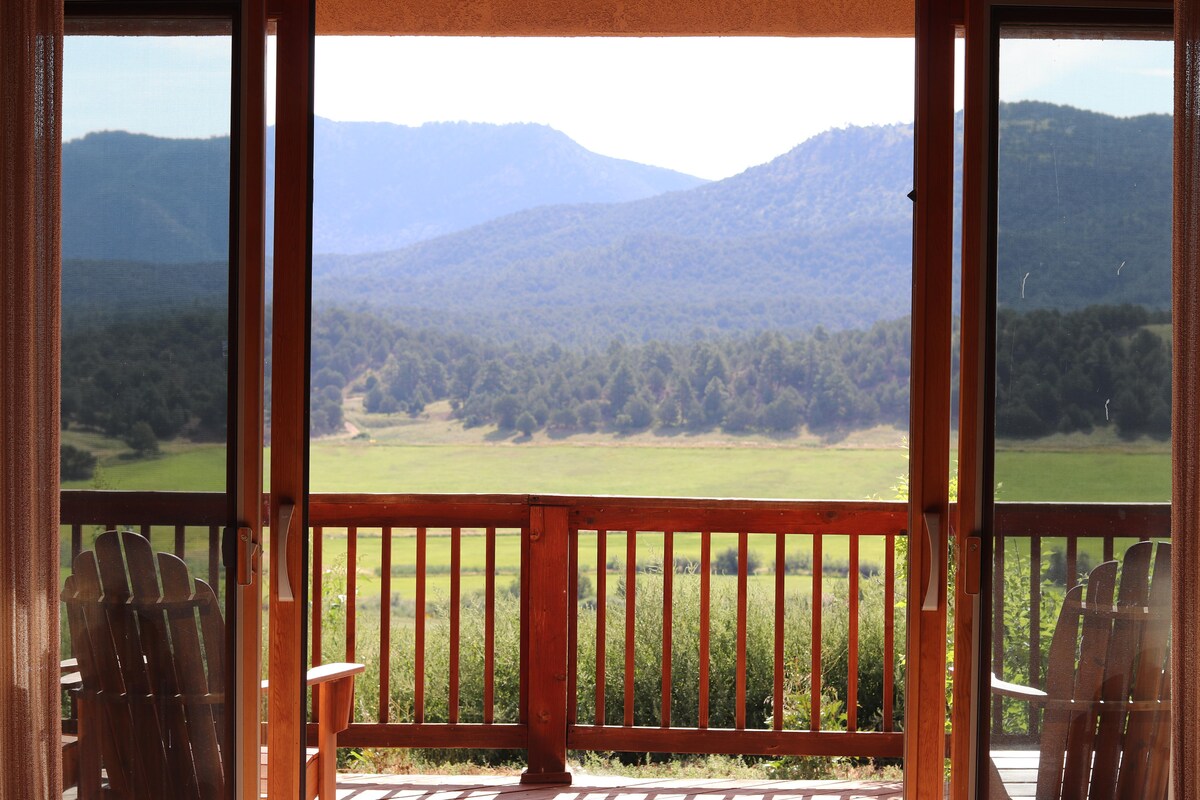A view from the porch captures rolling green hills and distant mountains under a clear sky. Two wooden chairs are positioned on a wooden deck, inviting relaxation. Natural light floods the space, enhancing the connection to the serene landscape.
