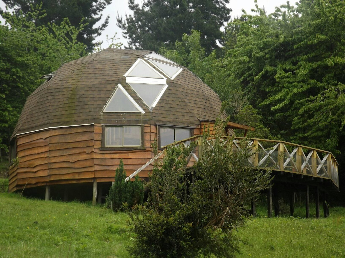 The dome-shaped cabin features a wooden exterior with large triangle-shaped windows, allowing natural light to fill the interior. A spacious deck surrounds the structure, blending with the landscape of grassy areas and trees nearby.