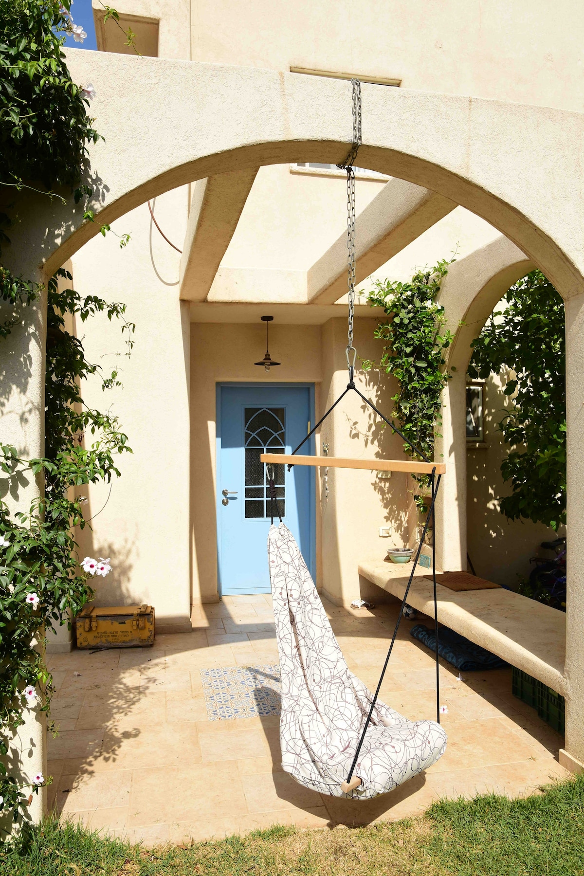 A charming entrance is framed by archways, featuring a swing chair suspended from a wooden beam. The light blue door and surrounding greenery contribute to a refreshing atmosphere, while the tiled floor extends to the outdoor seating area, inviting moments of relaxation.