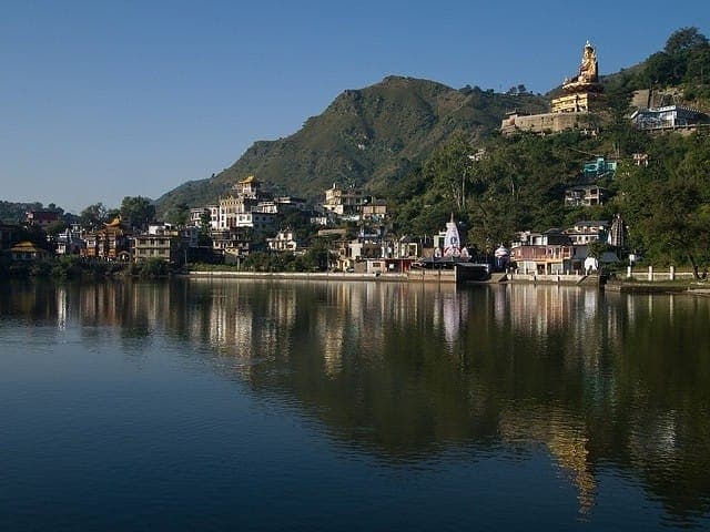 A serene landscape is captured, showcasing a tranquil body of water reflecting the hillside and buildings. Nestled among lush greenery, structures with distinct architecture are visible, along with a prominent statue overlooking the scene. The scene is bathed in natural sunlight, enhancing the calm atmosphere.