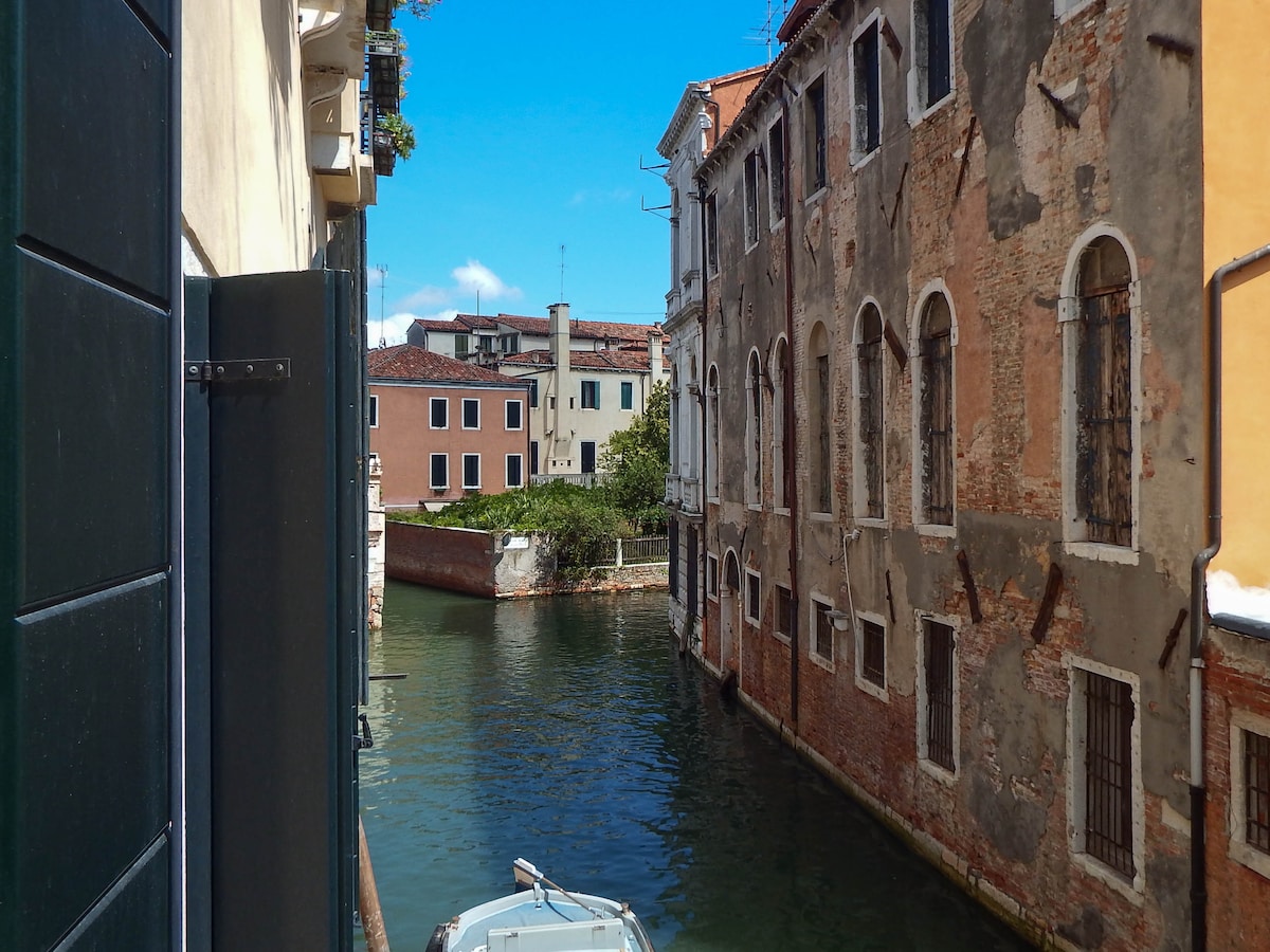 A view from a window reveals a narrow canal lined with weathered historic buildings. A small boat is seen gently floating on the water, while vibrant greenery peeks through the gaps. The sky is clear and blue, providing a bright backdrop to the scene.