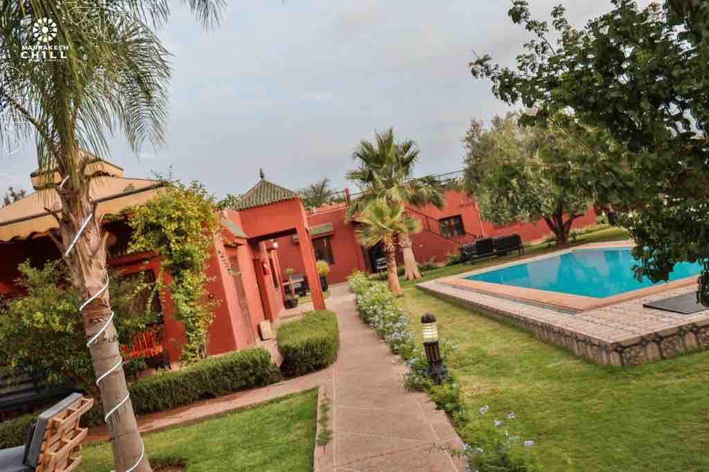 A well-maintained garden path leads to a turquoise pool surrounded by lush greenery and palm trees. The vibrant red exterior of the building is complemented by traditional architectural features, creating a serene outdoor environment ideal for relaxation.