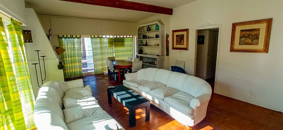 A bright living area features white sofas arranged around a central coffee table. Green patterned curtains frame large windows, allowing natural light to fill the space. A dining table can be seen in the background, complemented by shelves with decorative items.