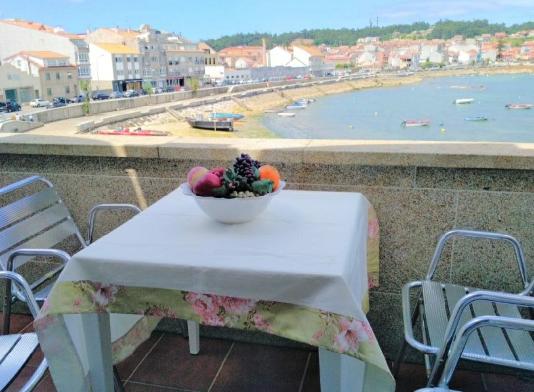 A terrace is shown with a small table covered in a floral tablecloth, featuring a bowl of colorful fruits. Metal chairs surround the table, and the view overlooks a scenic waterfront with boats and coastal buildings in the background.
