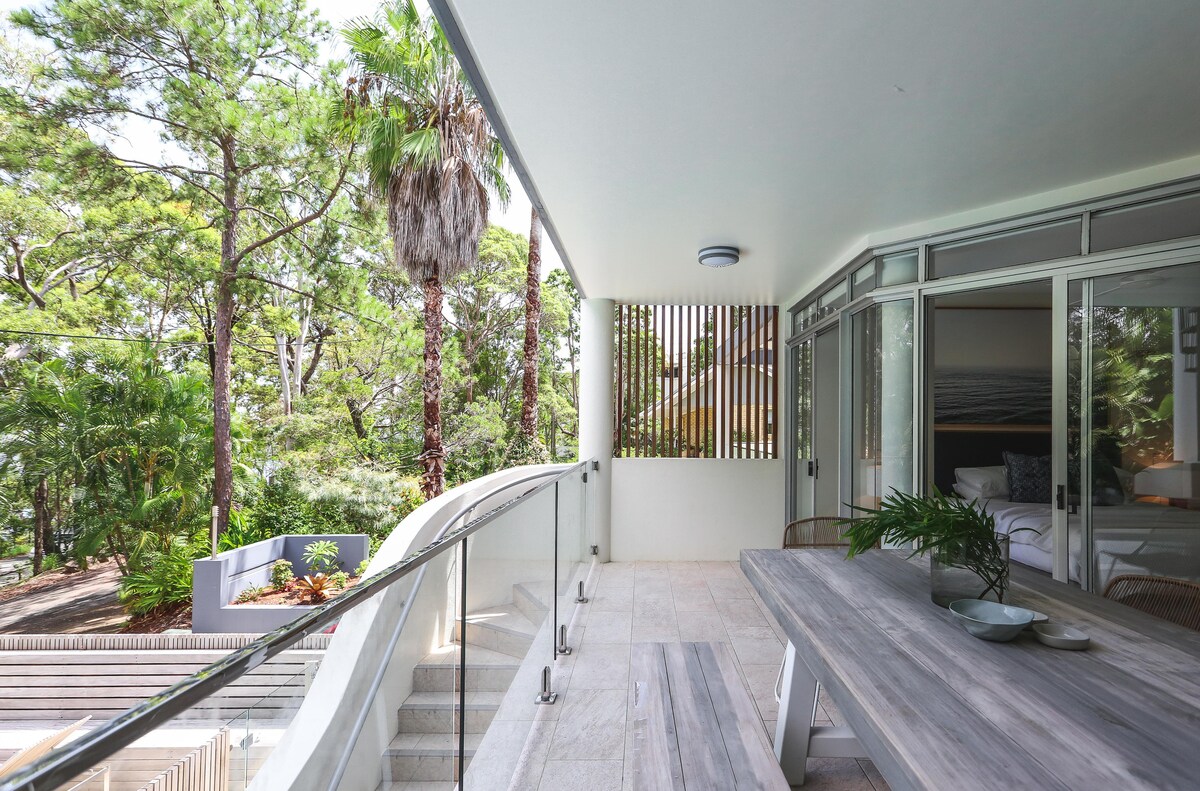 An outdoor terrace is presented with a long dining table complemented by a contemporary design. Surrounding greenery provides a sense of privacy, while glass railings enhance the view. The space is well-lit, inviting relaxation and outdoor dining amidst the natural landscape.