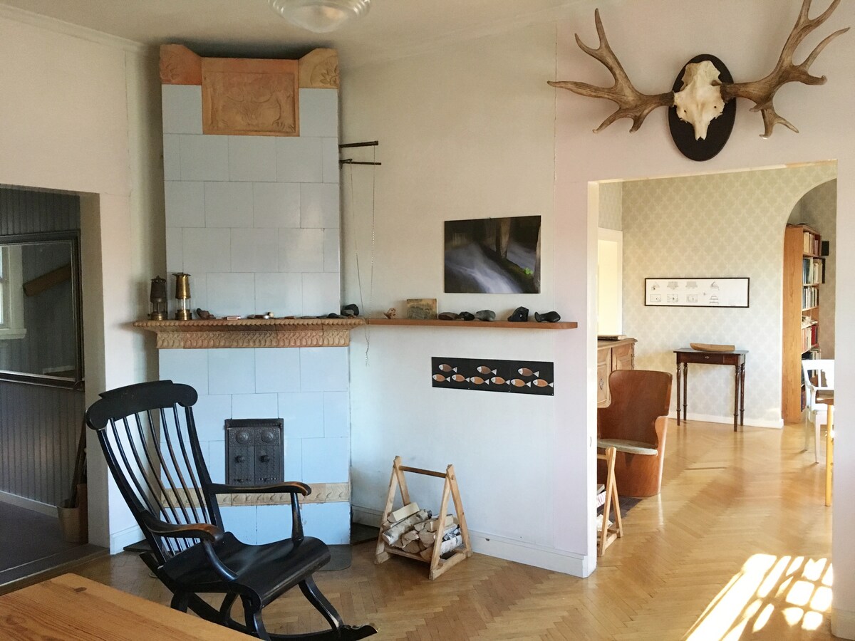 An inviting living space features a traditional tiled fireplace as the focal point, with a wooden rocking chair to one side. Decor includes an antler display above the fireplace, while light hardwood floors enhance the room's spaciousness. A glimpse of adjacent areas can be seen in the background.