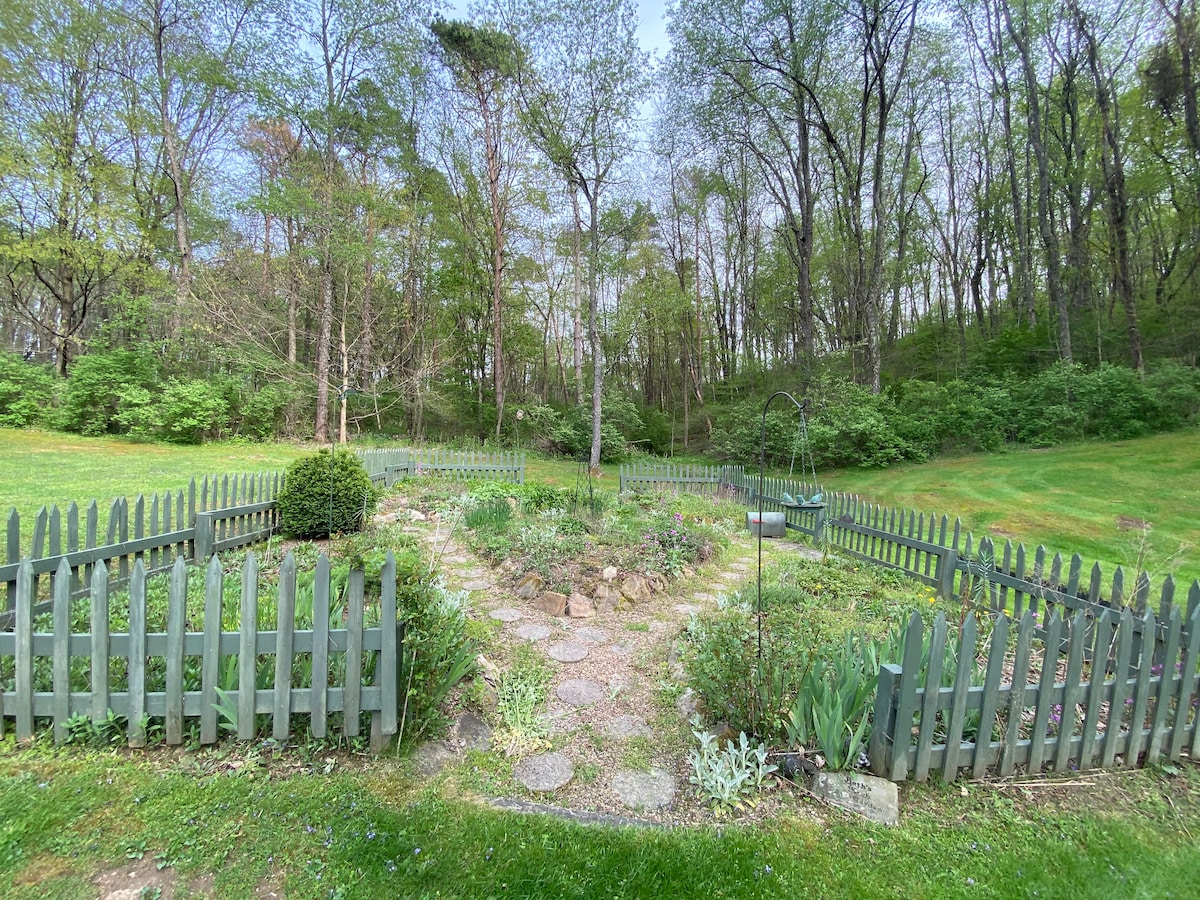 A lush garden is enclosed by a charming grey picket fence, featuring stone pathways that weave through various flowerbeds. Surrounding greenery consists of towering trees and vibrant plant life, creating a serene natural setting.