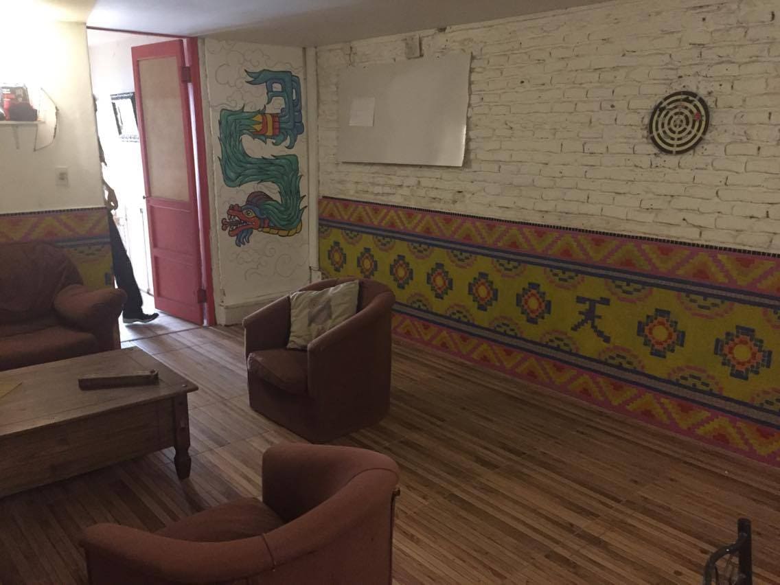 A communal living area features warm wooden flooring and colorful wall decorations. Two comfortable armchairs are positioned around a low wooden table. An artistic mural adds character to the space, while a red door leads to another area, inviting interaction and relaxation.