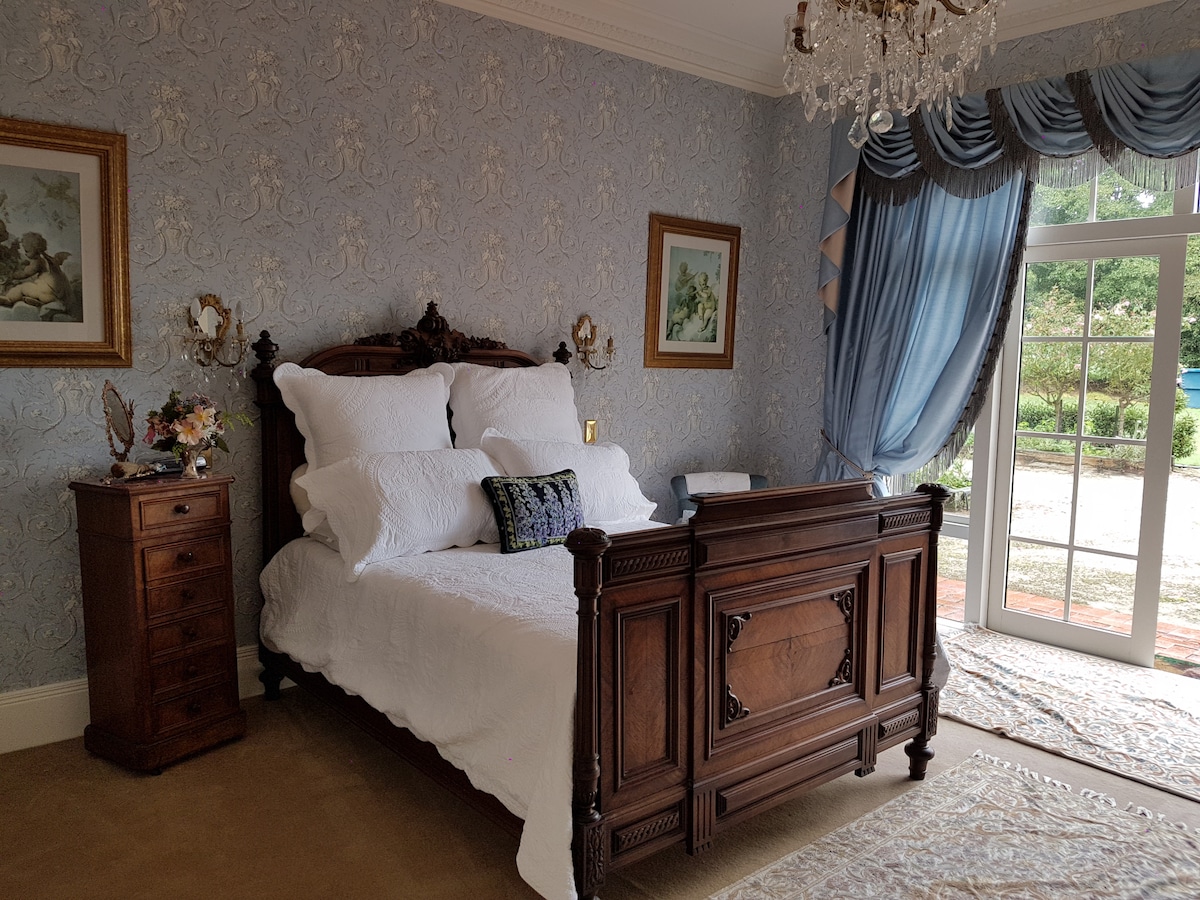 A spacious bedroom features an ornate wooden bed with plush white bedding. Decorative pillows add subtle color. Elegant blue drapes frame a large window, allowing natural light to fill the room. A charming wooden dresser is positioned nearby, enhancing the classic decor.