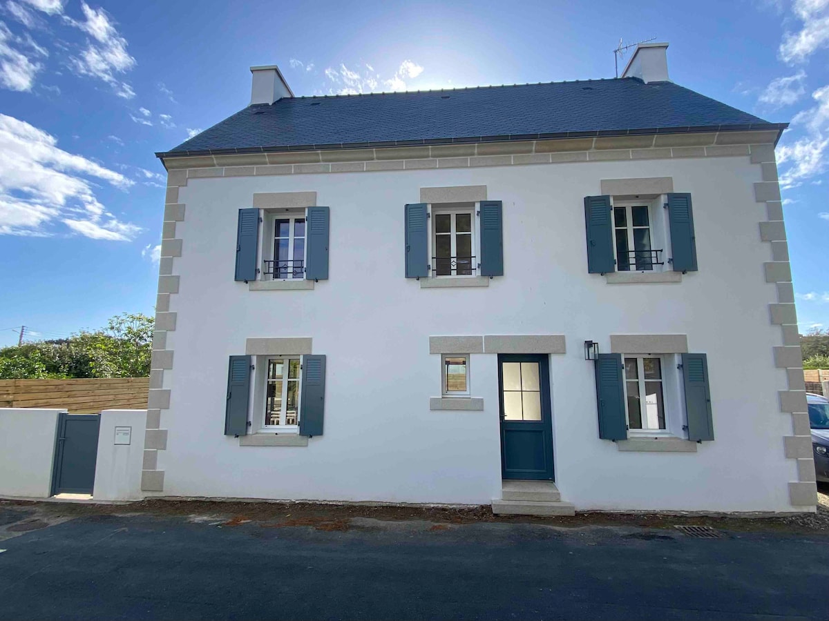 The exterior of a traditional Breton-style house is presented, featuring a white facade with dark blue shutters and a front door. The structure is two stories tall, with multiple windows allowing natural light. A quiet street view is visible, along with surrounding greenery.