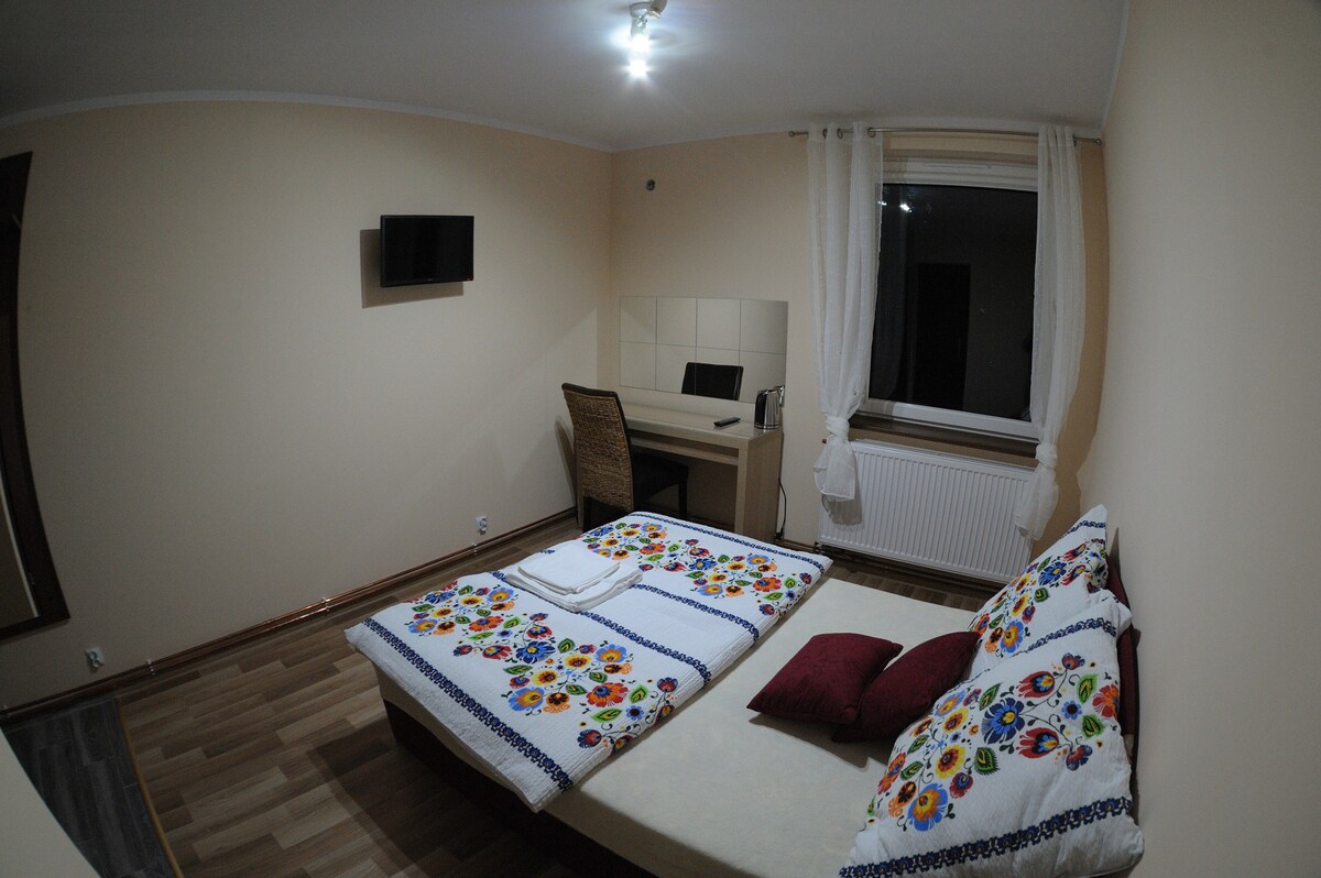 A simple bedroom features a spacious bed adorned with colorful, floral-patterned pillows and a neatly folded towel. A small desk with a chair is positioned near a large window, through which soft light enters. A flat-screen TV is mounted on the wall opposite.