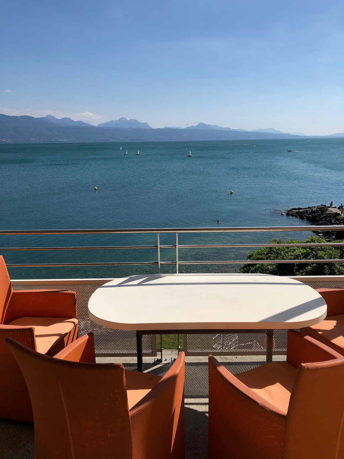 A scenic balcony features a white table surrounded by four orange chairs, overlooking a tranquil lake with sailboats in the distance. The view captures the natural beauty of the water and mountains under a clear blue sky.