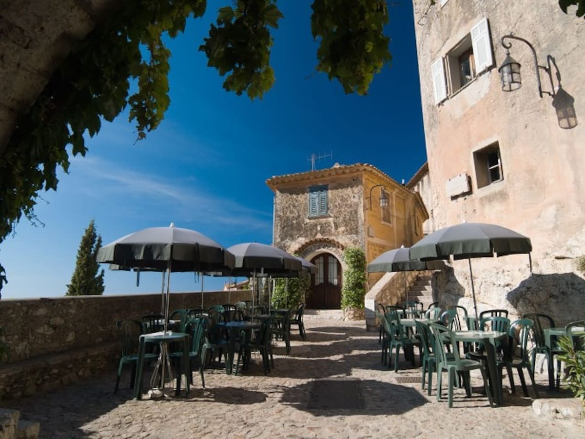 An outdoor seating area features several green tables and chairs beneath large gray umbrellas. The stone pathway leads to a historic building with a rustic facade, surrounded by green vines and trees under a clear blue sky.