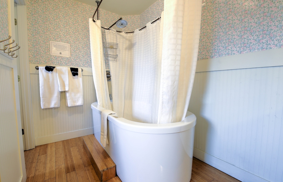 An open layout bathroom features a modern spa tub with a sleek, curved design. A showerhead is positioned above, and a white shower curtain encloses the space. Light bamboo flooring contrasts with floral wallpaper, while two plush towels hang on a wall-mounted rack nearby.