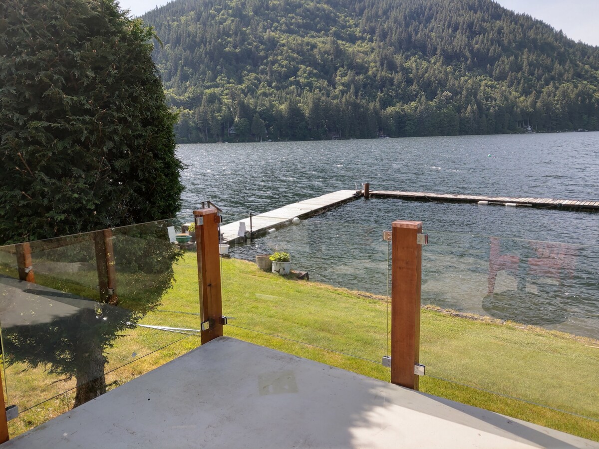 A scenic view of Lake Whatcom is presented from the deck, showcasing a large dock extending into the water. Lush greenery and mountains frame the landscape, while a glass railing offers an unobstructed sight of the tranquil setting.