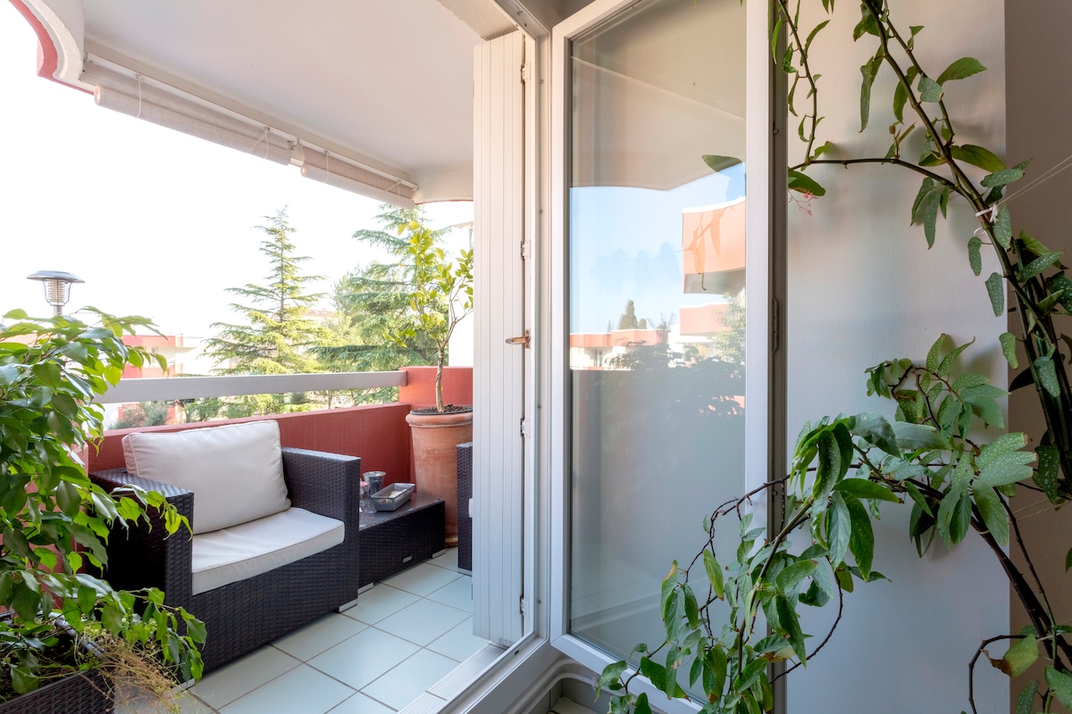 A small balcony is presented, featuring a rattan seating area with cushions. Lush greenery surrounds the space, while a tall potted plant adds height. A sliding door allows access to the indoor area, with a light breeze hinted by the soft foliage.