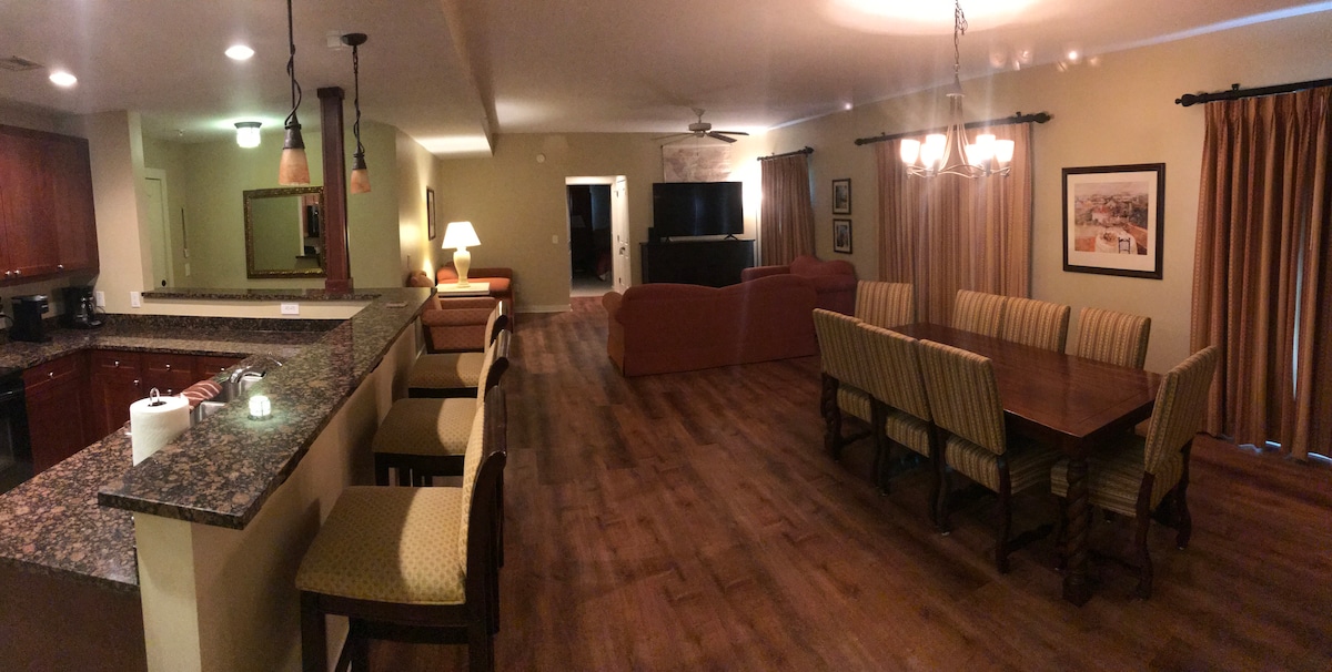A spacious living area is outlined by warm wooden floors. A dining table is flanked by cushioned chairs, while a granite countertop bar with stools offers additional seating. Soft lighting from overhead fixtures and lamps enhances the inviting atmosphere, with a large television visible in the background.