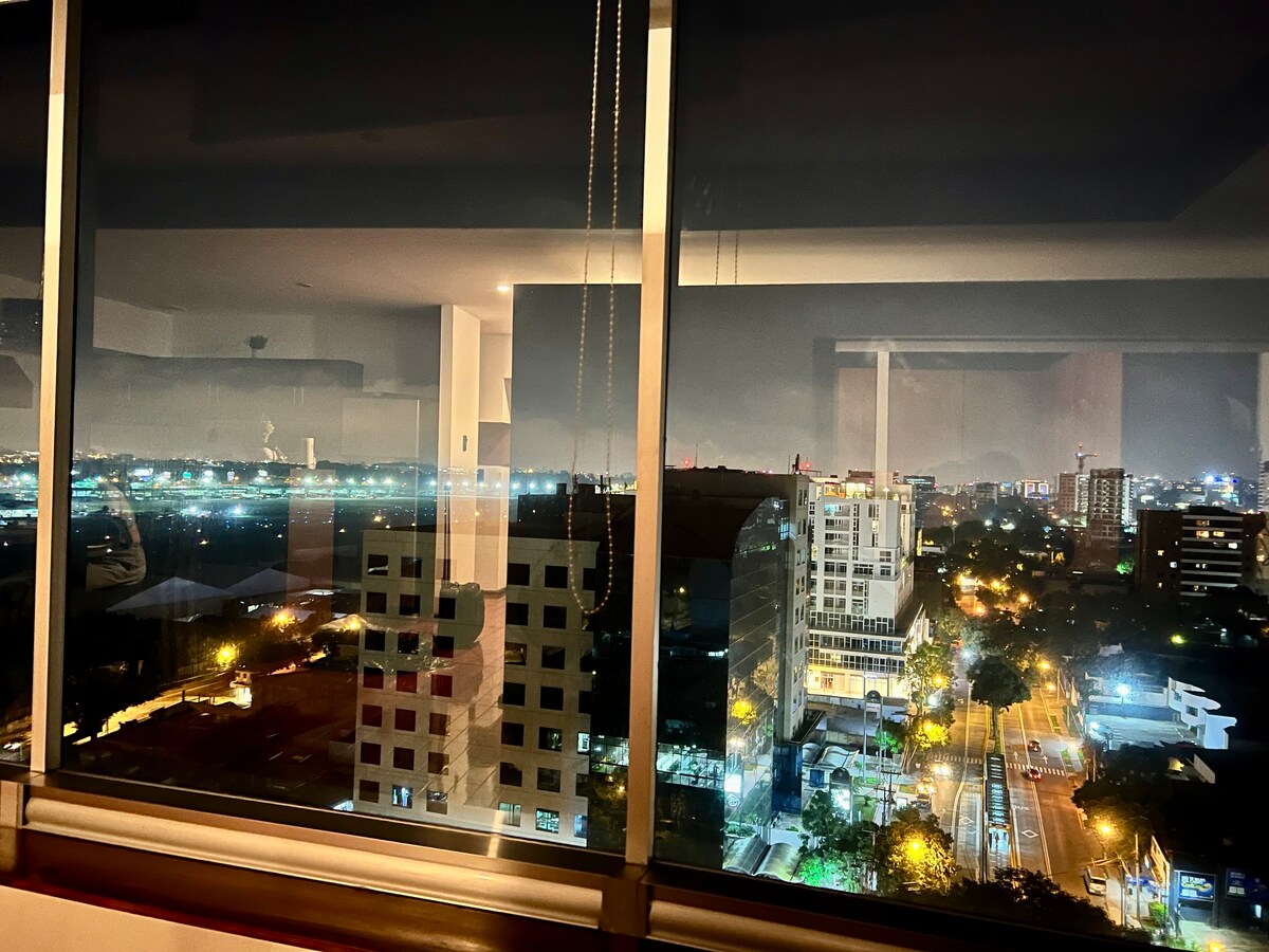 A city skyline is visible through expansive glass windows, revealing illuminated buildings and streets at night. The urban landscape is complemented by a distant view of lights twinkling across the horizon, creating a serene nighttime scene.