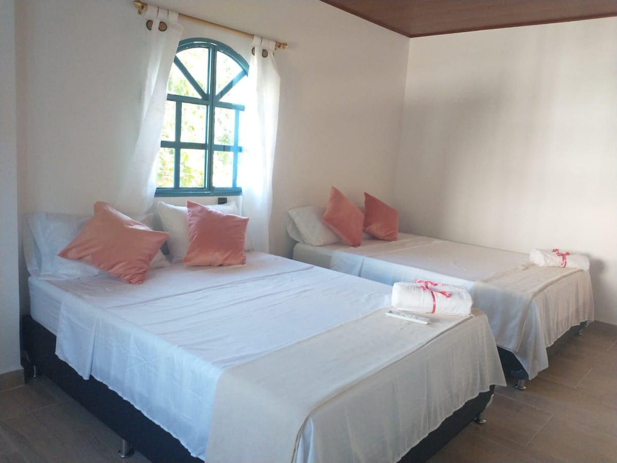 A well-lit sleeping area features two spacious beds adorned with crisp white linens and decorative cushions. The room showcases a charming arched window framed in blue, allowing natural light to illuminate the space. Fresh towels are neatly arranged at the foot of each bed.