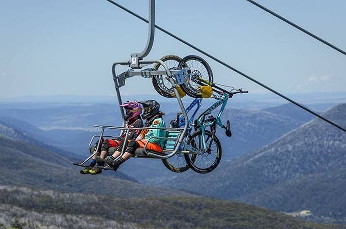 Two individuals are seated on a ski lift, with their mountain bikes suspended above them. The backdrop features rolling mountains under a clear sky, highlighting the region's natural landscape.