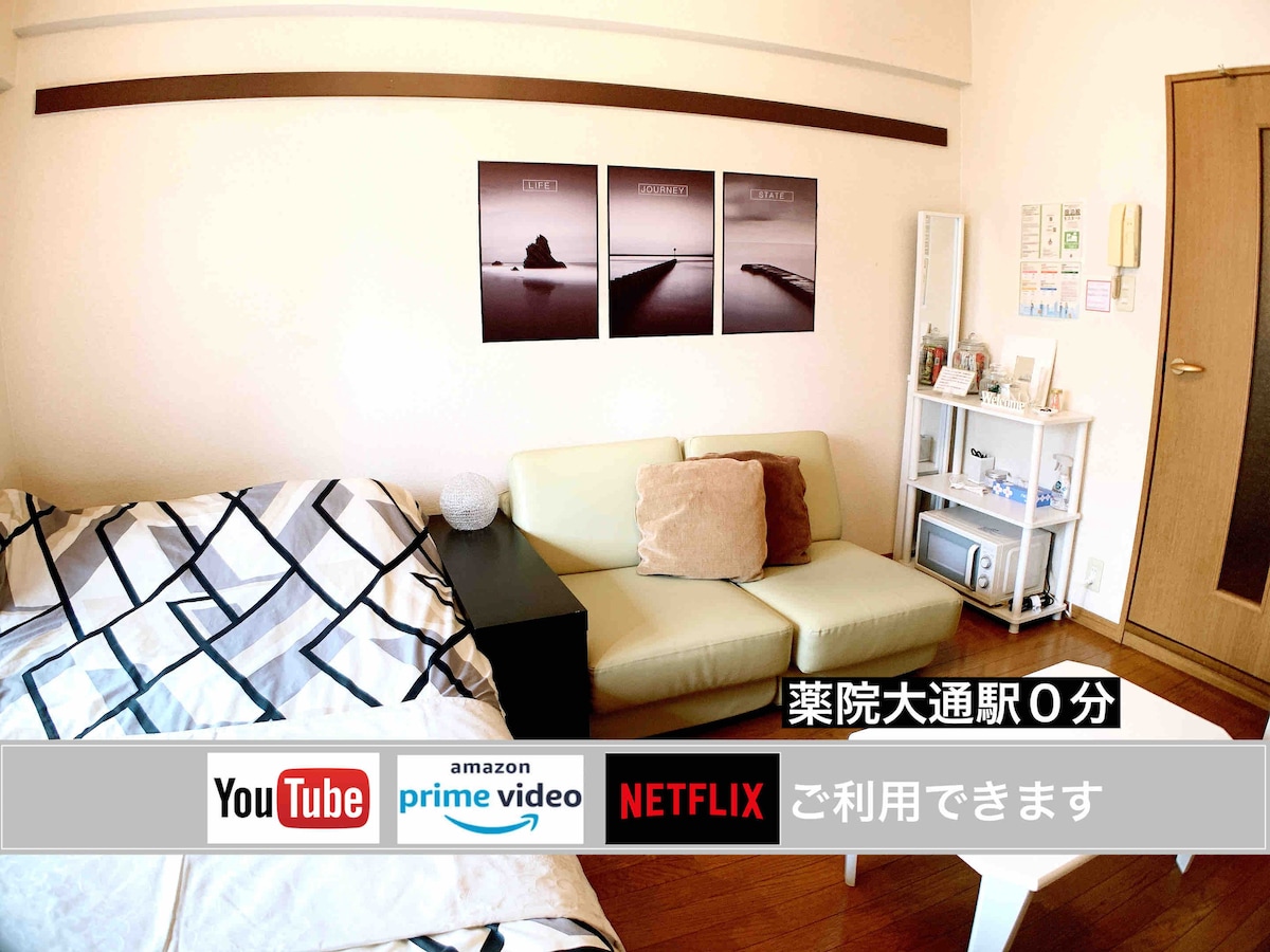 A compact room features a light beige sofa and an unmade bed with a patterned blanket. Three black and white framed photographs adorn the wall above the sofa. A small entertainment unit displays logos for streaming services, including Netflix and YouTube.