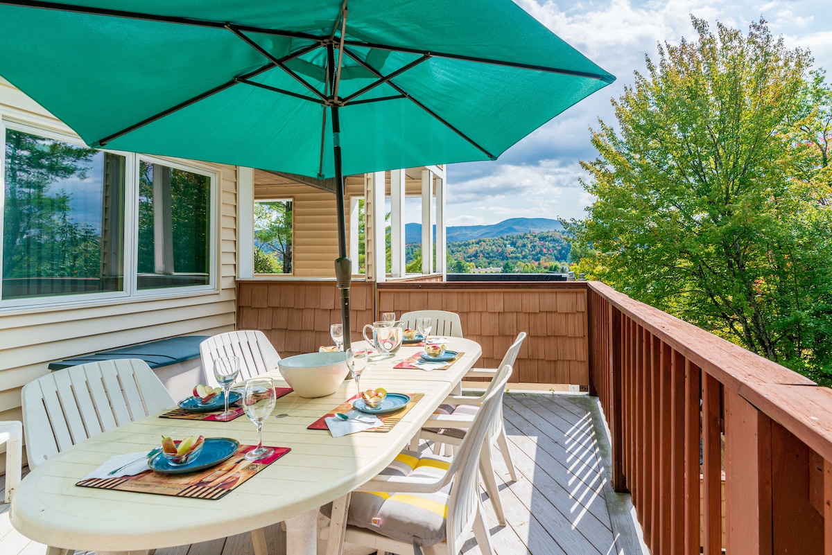 A spacious outdoor dining area is equipped with a large table set for six, featuring colorful plates and glasses. An oversized umbrella provides shade, with a scenic view of surrounding hills and trees visible in the background.