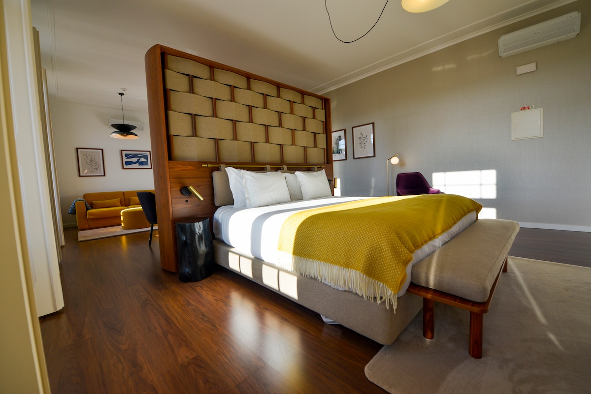 A spacious bedroom features a large, upholstered headboard and a neatly made bed with a warm yellow throw. Natural light filters through the window, illuminating the wooden floor and highlighting elegant furniture, including a cozy sitting area in the background.