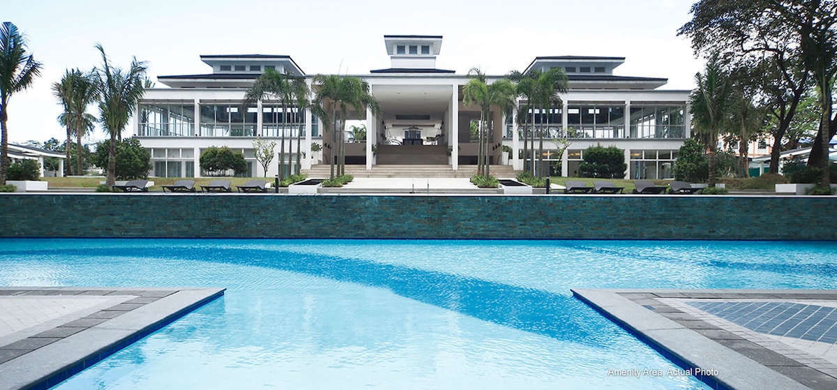 A large pool reflects the surrounding landscape, featuring vibrant greenery and modern architecture. The building in the background is spacious, with multiple levels and wide staircases leading to an airy entrance. Comfortable lounge chairs are arranged along the pool's edge, inviting relaxation.