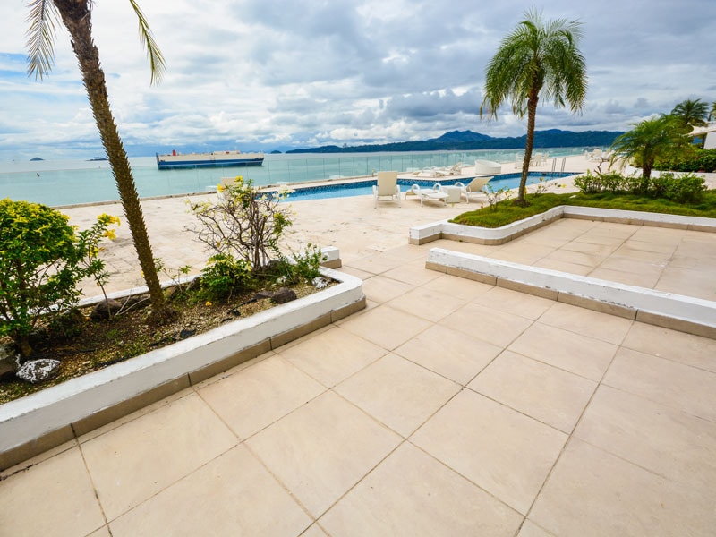 A wide outdoor space features a tiled patio leading towards a shimmering pool. Palm trees and vibrant yellow flowers are planted along the edge, framing a picturesque view of the ocean and distant mountains under a cloudy sky.