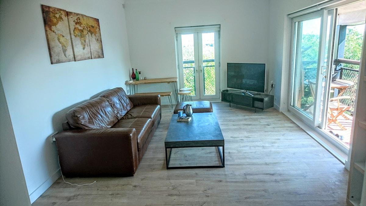 The living area features a large brown leather sofa accompanied by a coffee table. A television is positioned on a stand, with light entering through expansive glass doors that lead to a balcony. A wall-mounted world map artwork adds an element of design.
