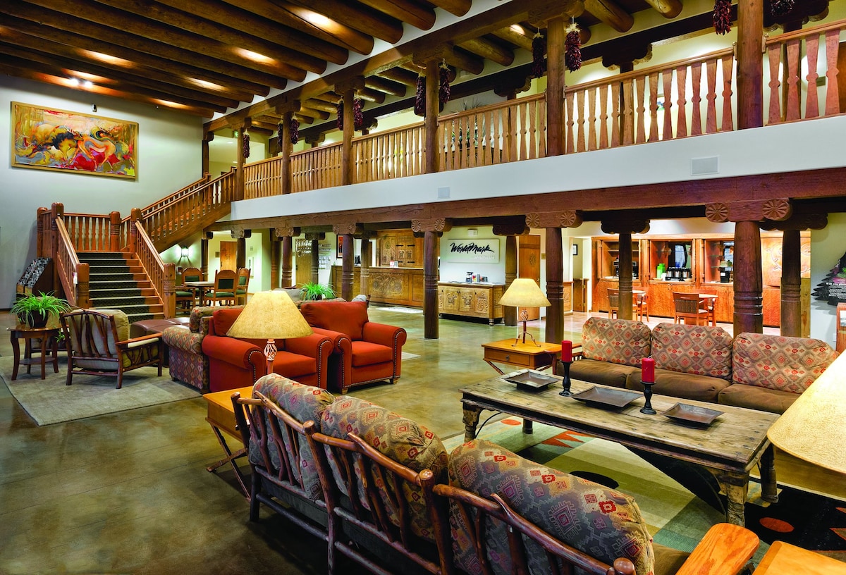 The spacious lobby features wooden beams and columns, creating a rustic ambiance. Comfortable seating areas are arranged around tables, complemented by decorative lamps. A staircase leads to the upper level, while a front desk area is visible in the background.