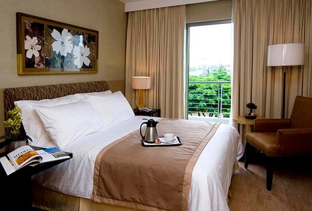 A well-appointed room features a spacious bed with crisp white linens and a light brown throw. A coffee and tea set is placed on a small table beside the bed, and a comfortable chair is positioned nearby. Natural light enters through a large window adorned with light curtains.