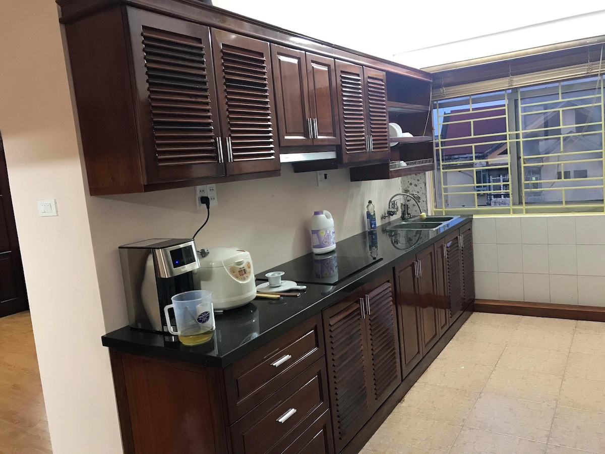 A functional kitchen is presented with wooden cabinetry and a dark countertop. Appliances such as a coffee maker and rice cooker are visible. Natural light streams in through a window fitted with light-colored blinds, highlighting the organized layout and available counter space.