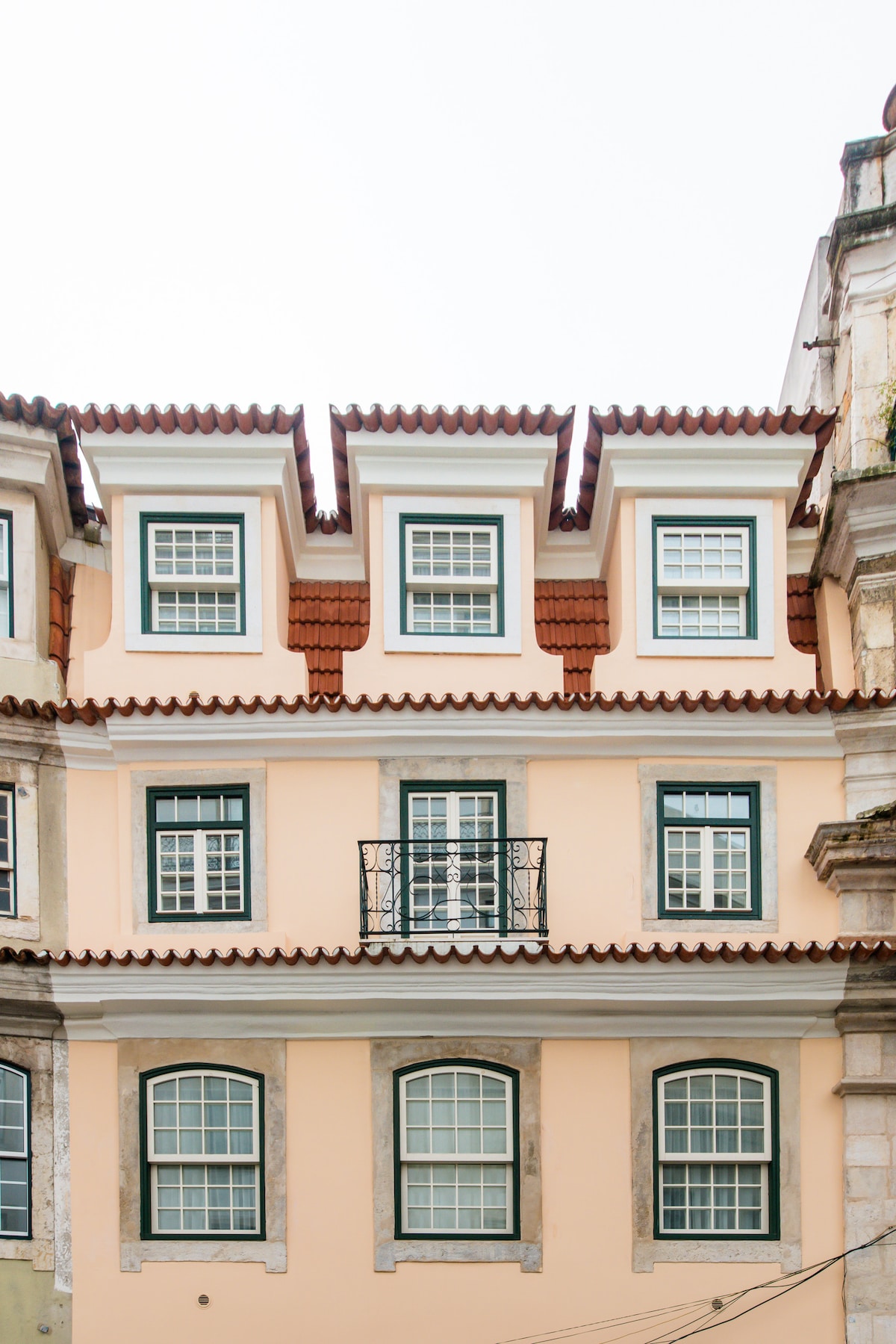 The exterior of a traditional building is shown, featuring a soft peach facade and a distinct tiled roof. Multiple windows with green shutters are visible, complemented by a small wrought-iron balcony, adding to the architectural charm of the structure.