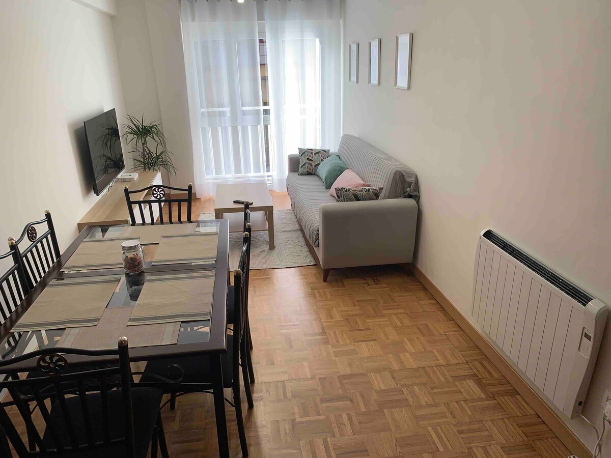 A bright living area features a comfortable sofa, a dining table with seating for six, and a Smart TV. Natural light enters through large windows dressed with sheer curtains, and wooden flooring provides a warm foundation. Simple wall art decorates the space.