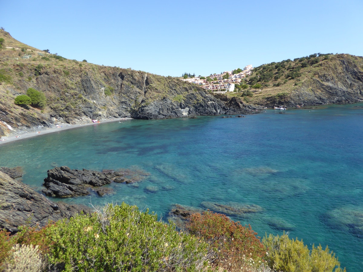 A rocky coastline is depicted, with clear blue waters gently lapping at the shore. The beach can be seen in the foreground, surrounded by lush greenery and rugged cliffs. In the distance, a quaint village is nestled on a hillside, adding to the serene coastal landscape.