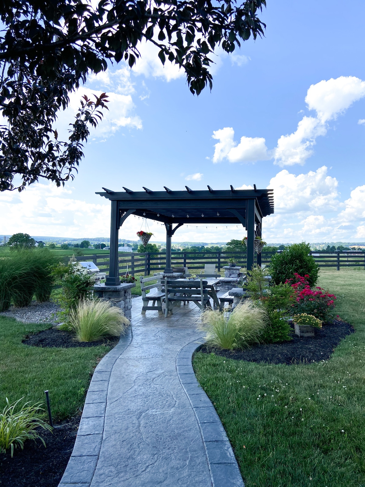 A stone pathway leads to a shaded outdoor seating area, framed by lush greenery and colorful flower beds. The pergola above, adorned with hanging plants, provides a tranquil space overlooking open farmland and distant rolling hills under a bright blue sky.