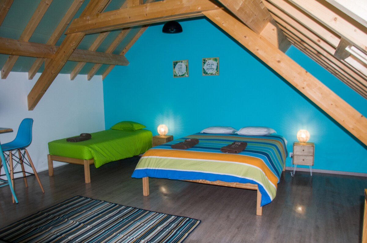 An attic space features two beds, one with a colorful striped cover and the other with a solid green blanket. Simple nightstands with lamps provide soft lighting. Exposed wooden beams and a blue accent wall create a relaxed ambiance, complemented by a striped area rug.