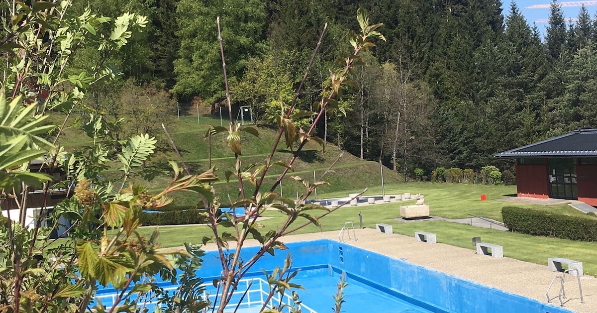 A serene outdoor swimming pool is framed by lush greenery and rolling hills. Surrounding areas include well-maintained lawns and a building with a dark roof, set against a backdrop of dense trees, creating a peaceful environment.
