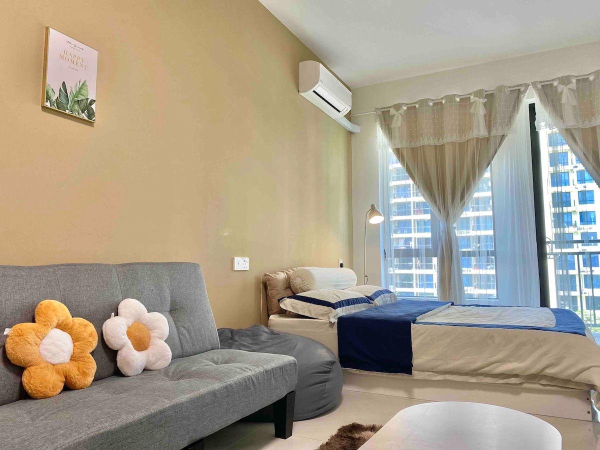 A cozy living area features a queen size bed with bedding in soft tones. A compact sofa bed and two bean bag chairs provide additional seating. Natural light floods the space through large windows adorned with sheer curtains, revealing a view of the surrounding cityscape.