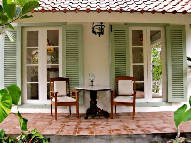 A quaint outdoor seating area features two wooden chairs beside a round table, framed by lush greenery. Large, white-framed windows with shutters offer a view into the villa, while a warm light fixture hangs overhead, contributing to the inviting ambiance.