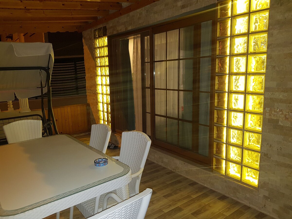An outdoor seating area features a table surrounded by four white chairs. Glass block walls provide ambient lighting, illuminating the space. A swing chair is observed in the corner, and wooden beams run overhead, enhancing the structure's natural feel.