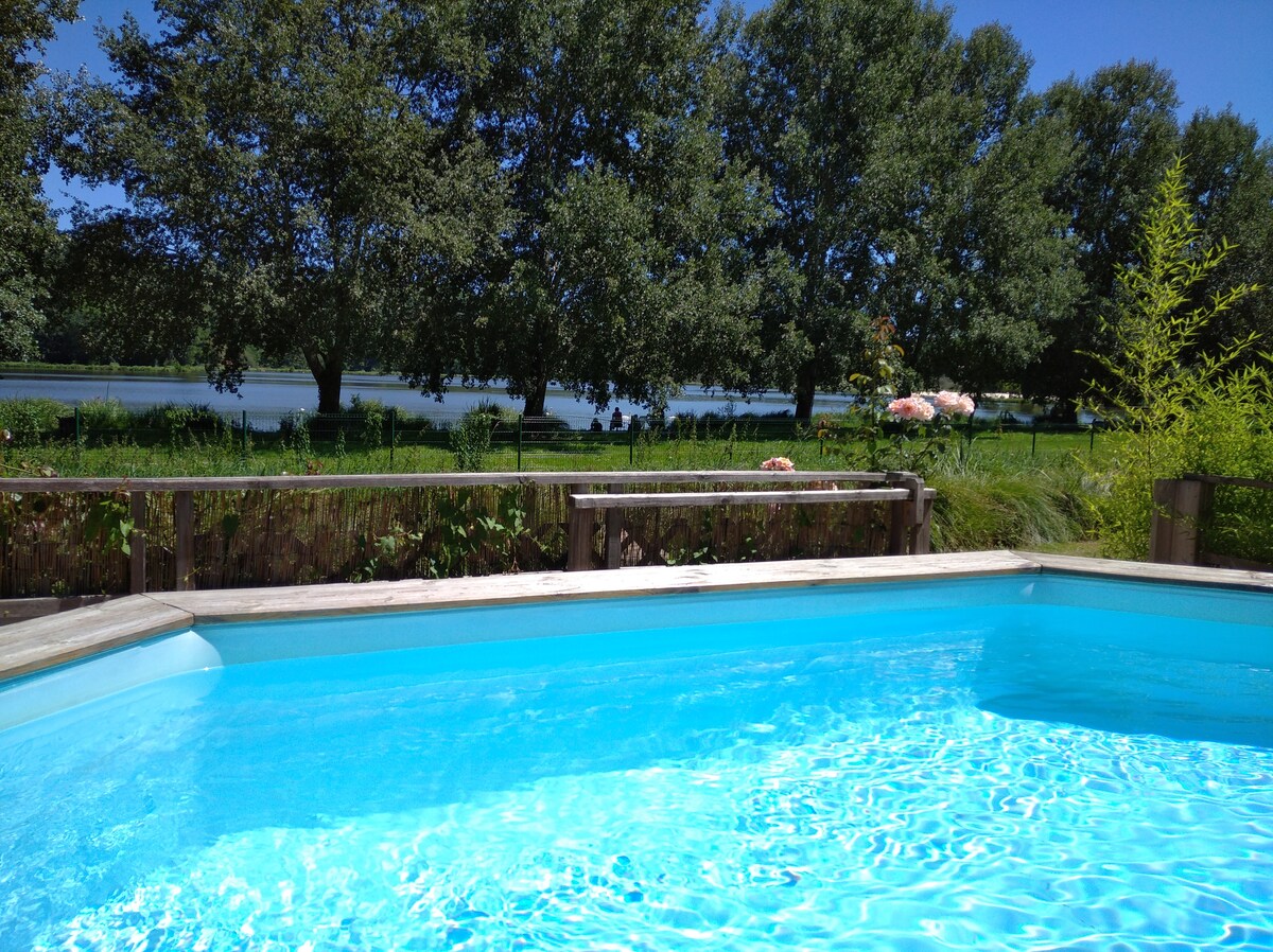 A private heated pool is depicted with clear blue water in the foreground, framed by a wooden deck. Lush greenery and several trees create a serene backdrop, leading to a calm lake visible in the distance under a bright sky.