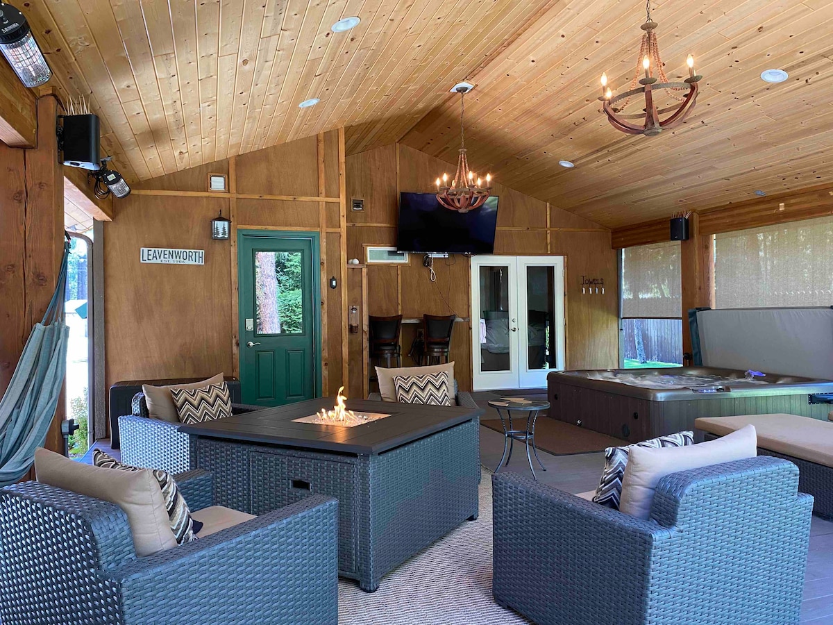 The indoor lounge area features a cozy seating arrangement with wicker chairs surrounding a fire table. A television is mounted on the wall, and a hot tub is positioned nearby. Natural wood accents and warm lighting create a welcoming ambiance.