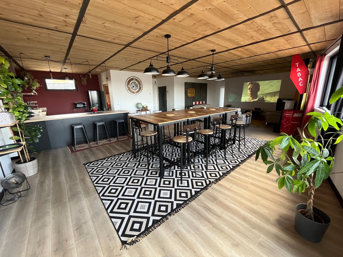 A spacious living area features a large wooden dining table surrounded by black chairs. An area rug with geometric patterns adds contrast to the light wooden floor. Plants are positioned around the space, and a well-equipped kitchen is visible in the background.
