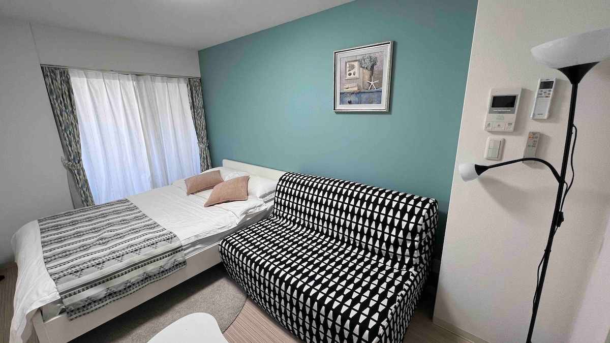A well-lit bedroom features a spacious bed with a light-colored duvet and two decorative pillows. A patterned sofa adds contrast and visual interest. The room is equipped with large curtains allowing natural light to filter in, and a framed artwork is displayed on the wall.