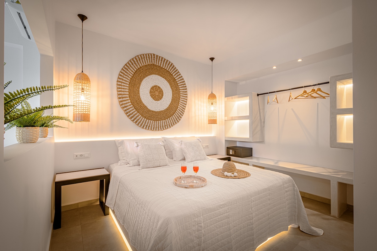 A well-lit bedroom features a spacious bed adorned with a light-colored quilt and decorative pillows. Stylish woven wall decor complements the space, with pendant lights hanging on either side. A small table holds two drinks, and a closet area provides hangers and storage.