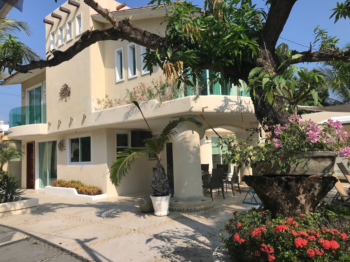 A two-story villa is surrounded by lush vegetation, featuring a blend of modern architecture and natural elements. Large windows and a spacious terrace provide views of the garden. Vibrant flowers and tropical plants enhance the serene outdoor environment, inviting relaxation.