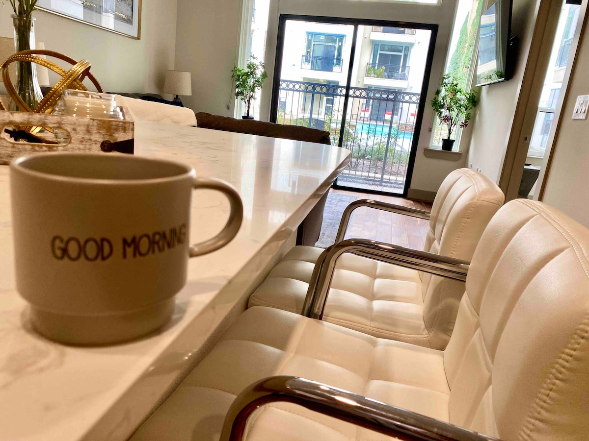 A sleek countertop is displayed with a coffee mug featuring the words 'GOOD MORNING.' Two white, modern chairs are positioned nearby, providing a view of the outdoor pool area through glass doors framed by potted plants.